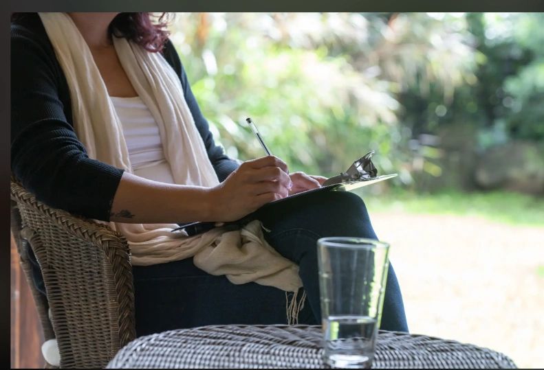 Person writing notes on a clipboard outdoors with a glass of water nearby.
