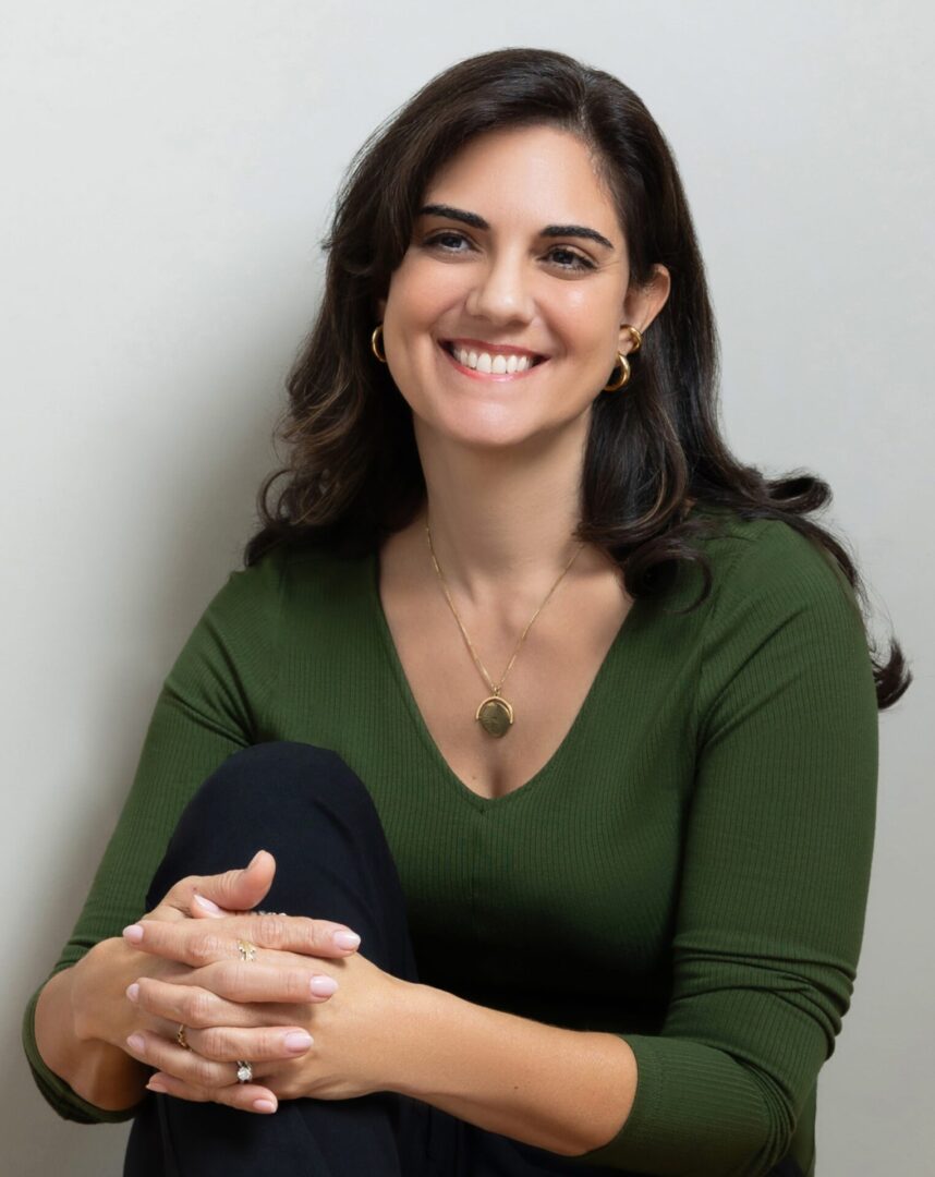 Smiling woman in green top sitting with hands folded, looking at camera.