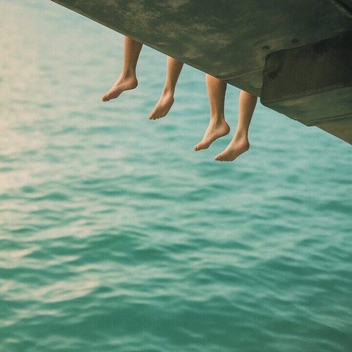 Three pairs of feet dangling above calm water from a dock.